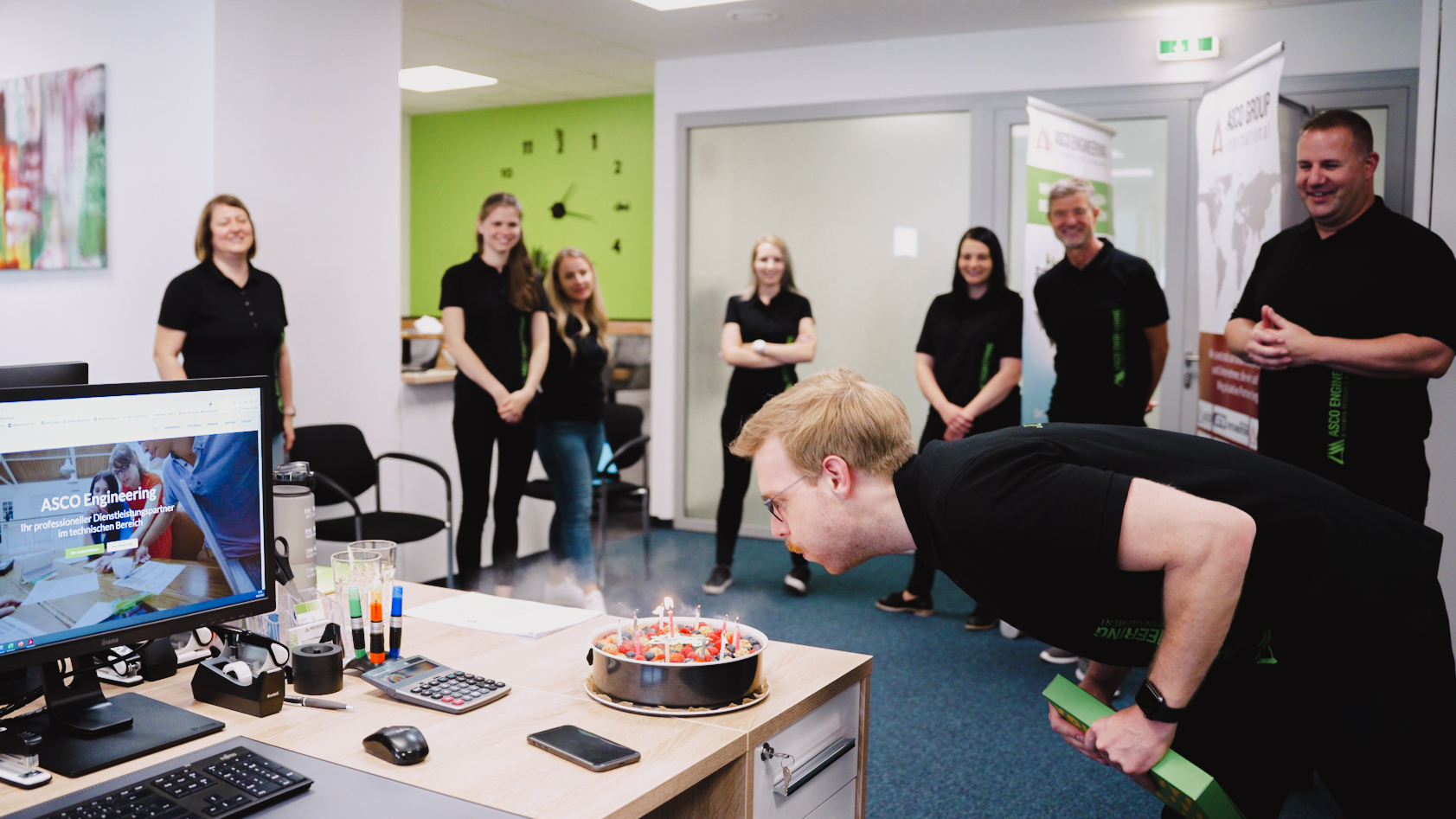 Gemeinsam feiern – Geburtstagsmoment im Team Eine Person pustet Kerzen auf einem Geburtstagskuchen aus, andere stehen lächelnd daneben.